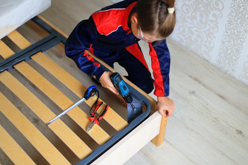 The guy fixes the bed frame with a drill and a screwdriver